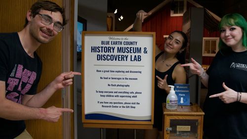Three Minnesota State Mankato Department of Art and Design students posing next to the welcome to the Blue Earth County History Museum and Discovery Lab entrance sign at the Blue Earth County Historical Society museum