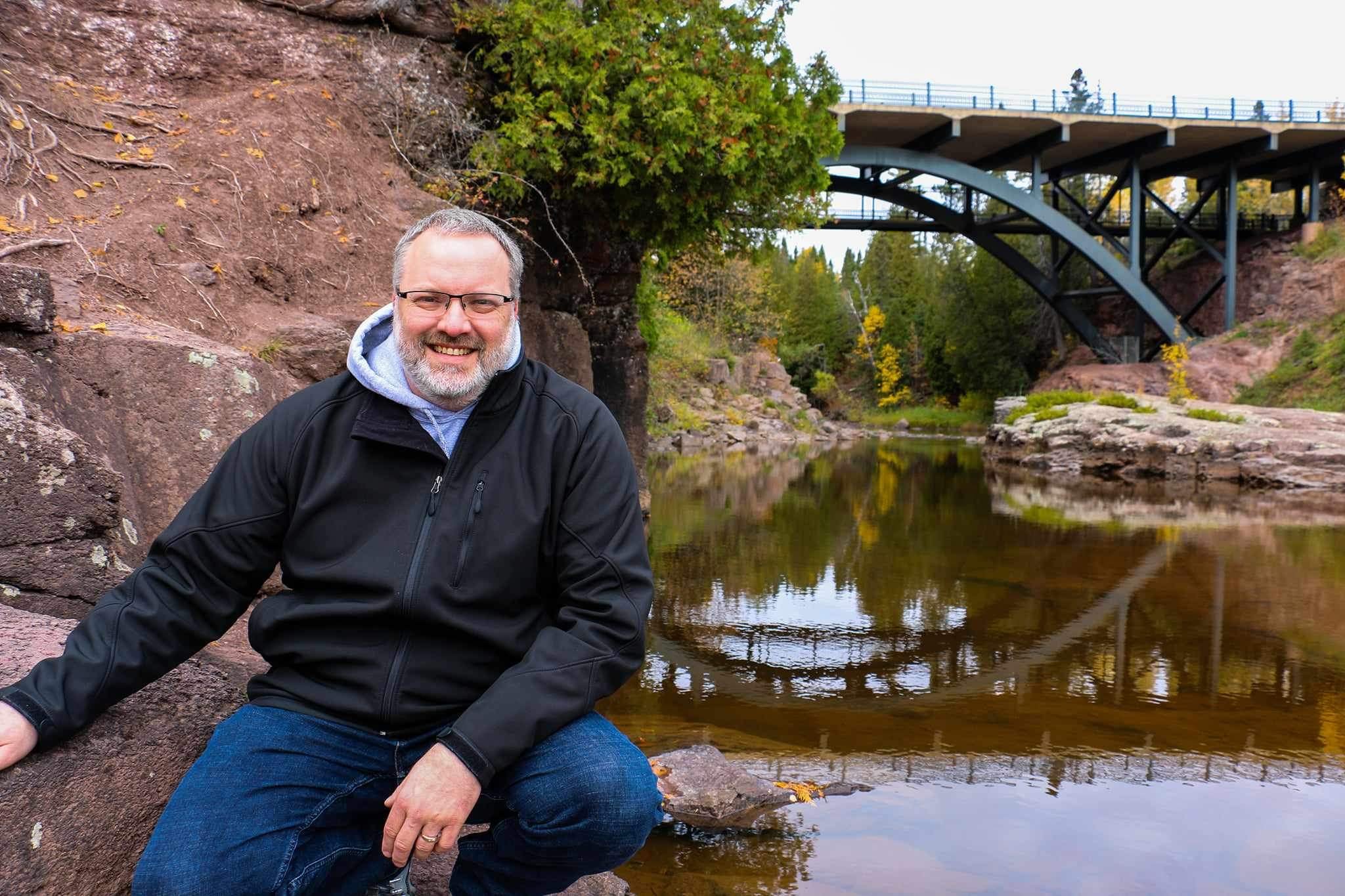 Man sitting next to river with bridge above.