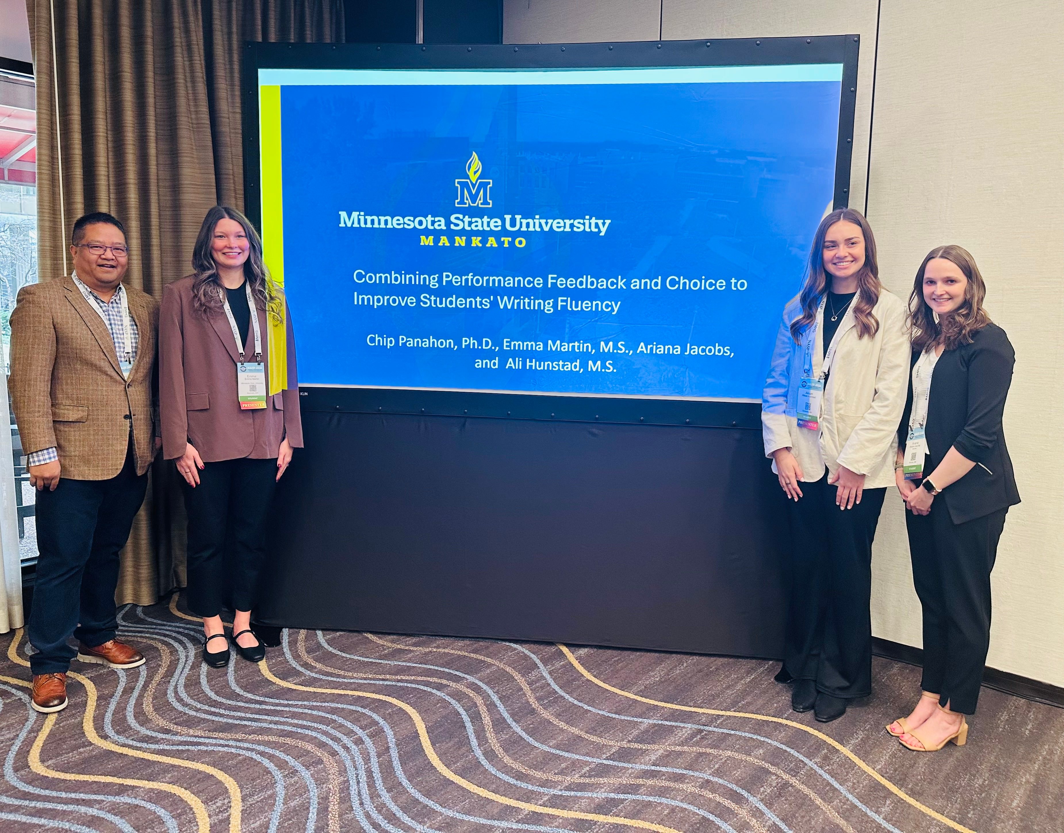 Four presenters stand beside a Minnesota State University, Mankato presentation on improving students&rsquo; writing fluency, smiling at a conference.