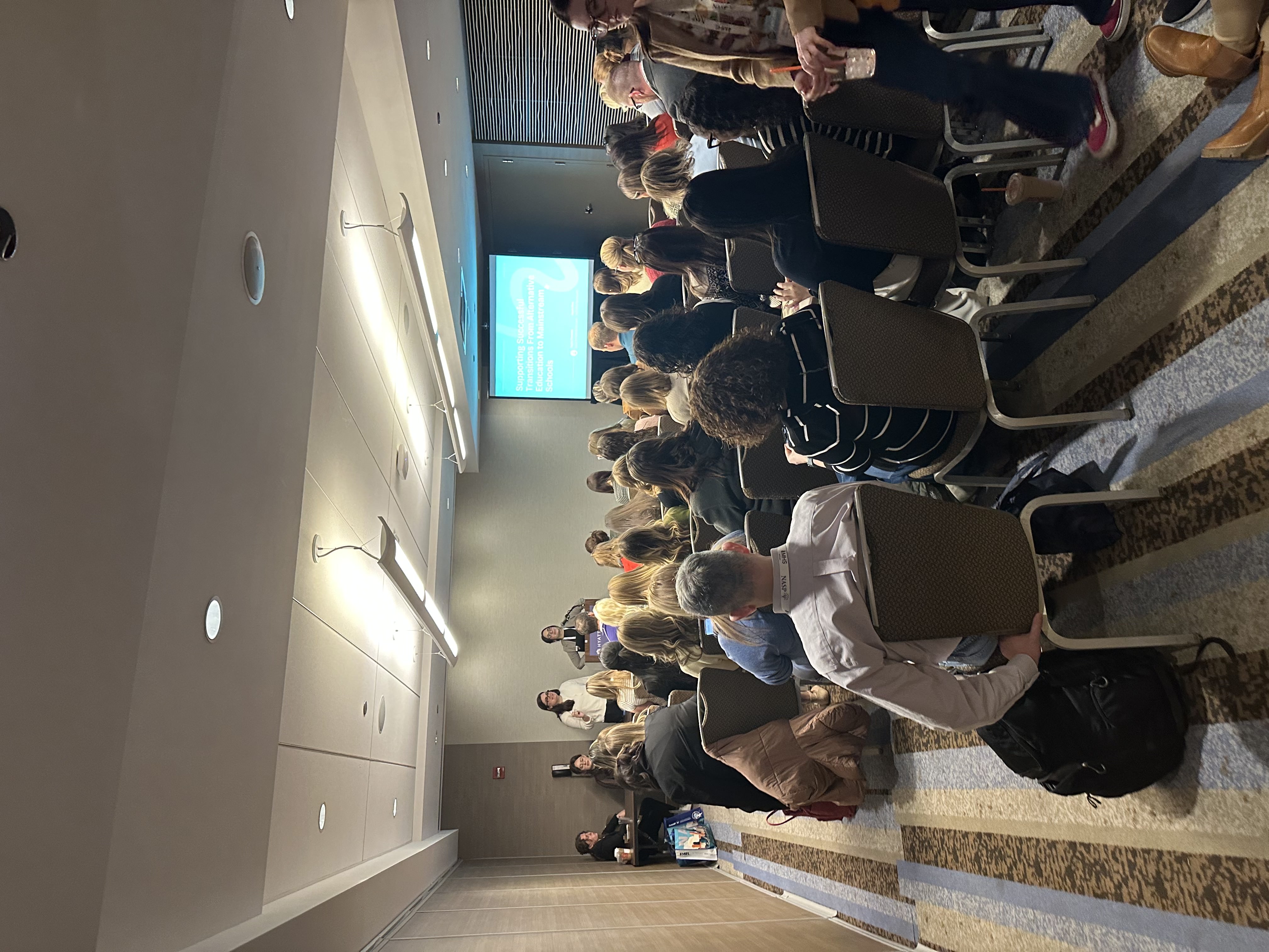 A conference room filled with attendees seated and listening to two presenters speaking at the front beside a projected presentation slide