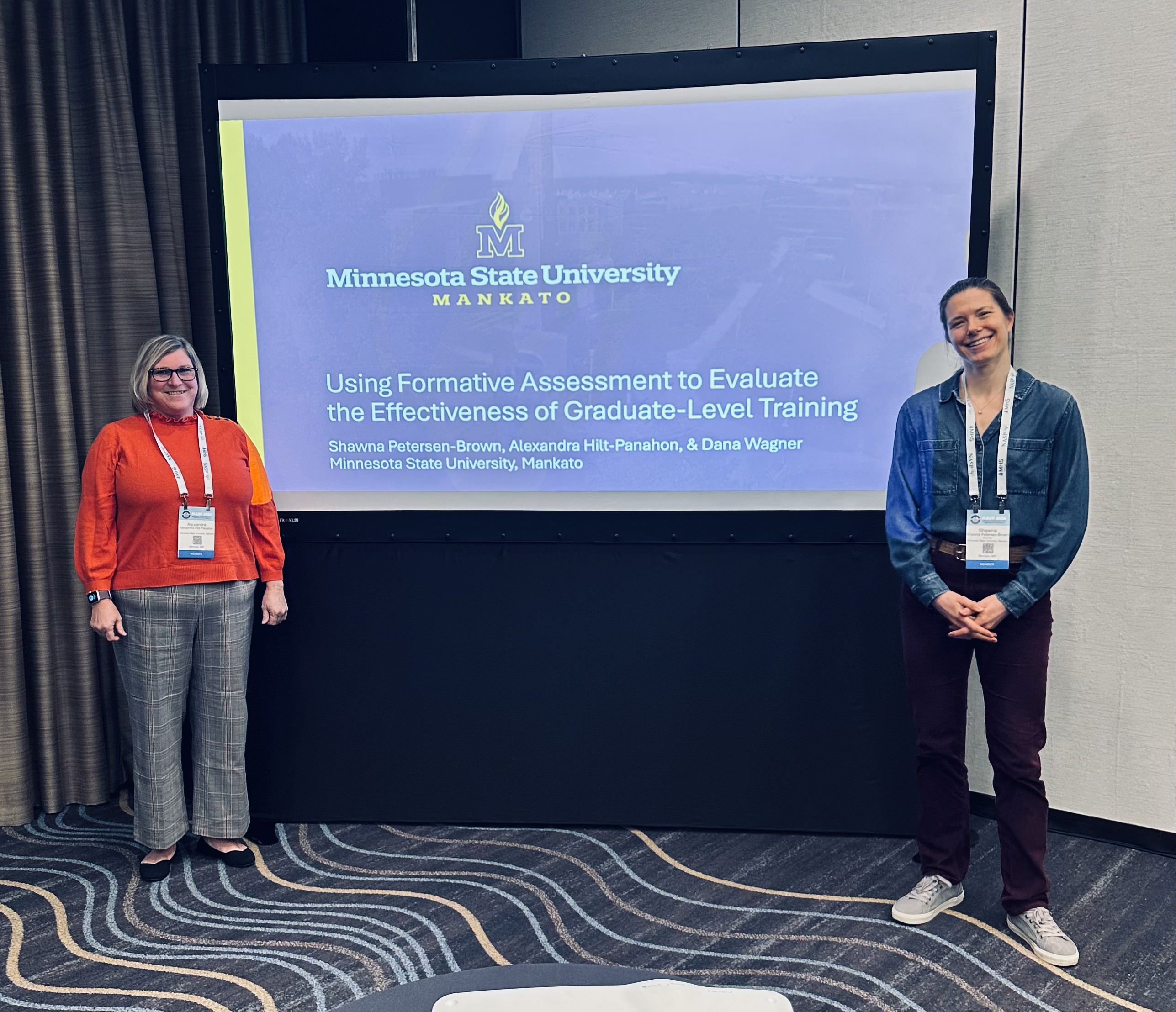 Two presenters stand beside a Minnesota State University, Mankato slide on evaluating graduate-level training, smiling at a conference