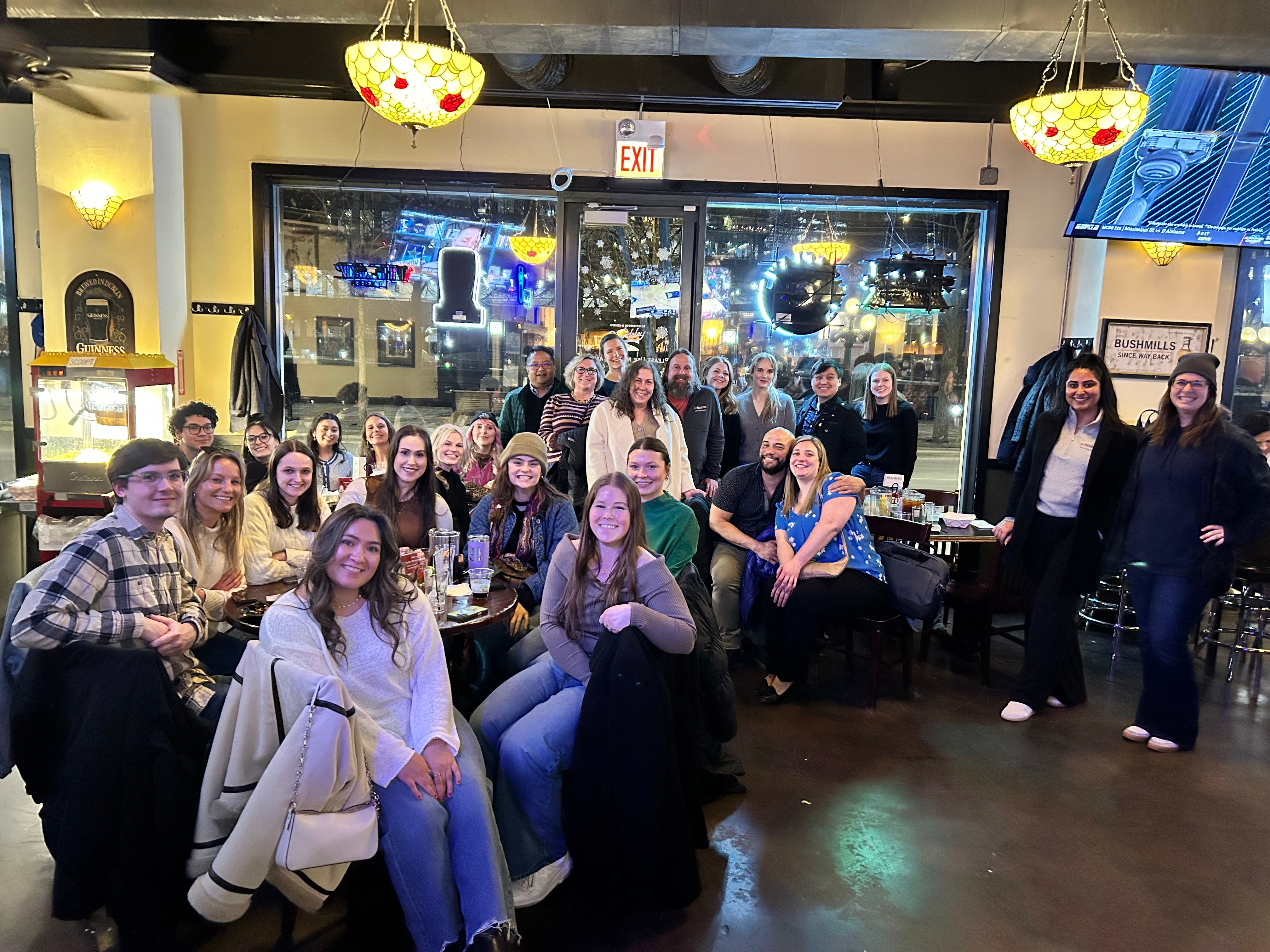 A large group gathers indoors around tables, smiling for a group photo