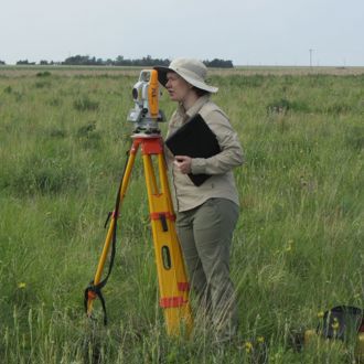 Alyssa Sims (Edwards) standing in a field and looking through survey equipment on a stand