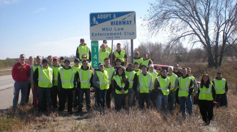 The Law Enforcement Club members posing as a group on the side of the road under a sign that says "Adopt a highway MSU Law Enforcement Club since 1995"