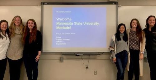 I-O psych students posing in the classroom next to the projection board that says welcome to Minnesota State University, Mankato