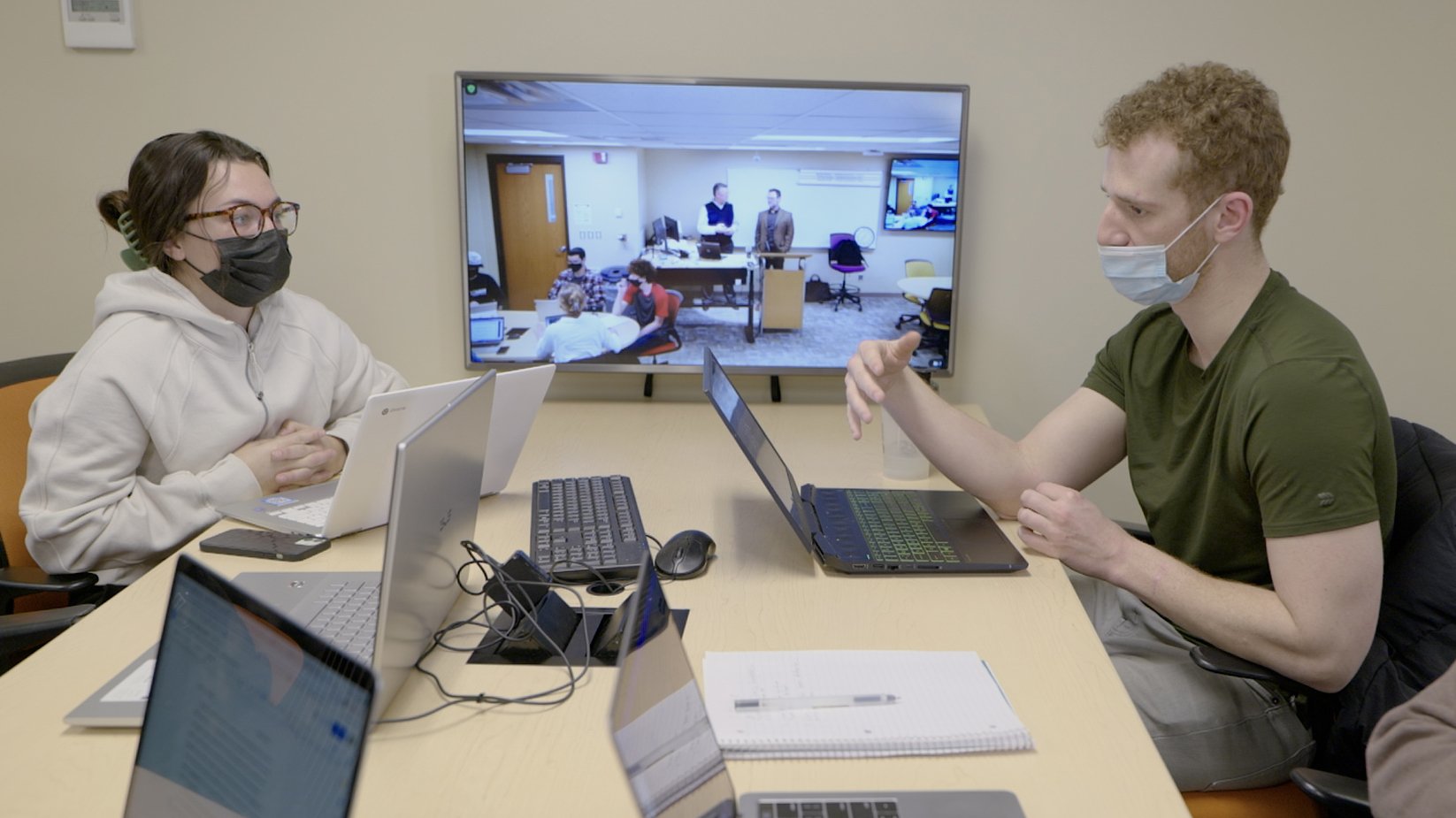 a group of people sitting at a table with laptops and a video conference